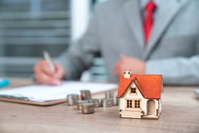 A person wearing a suit and tie signs a document at a desk, with coins and a house figurine in the foreground.