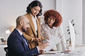 Three people stand over a desk reviewing paperwork in an office.