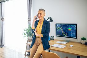 Woman leaning on a desk with her back to a computer monitor, talking on the phone. 