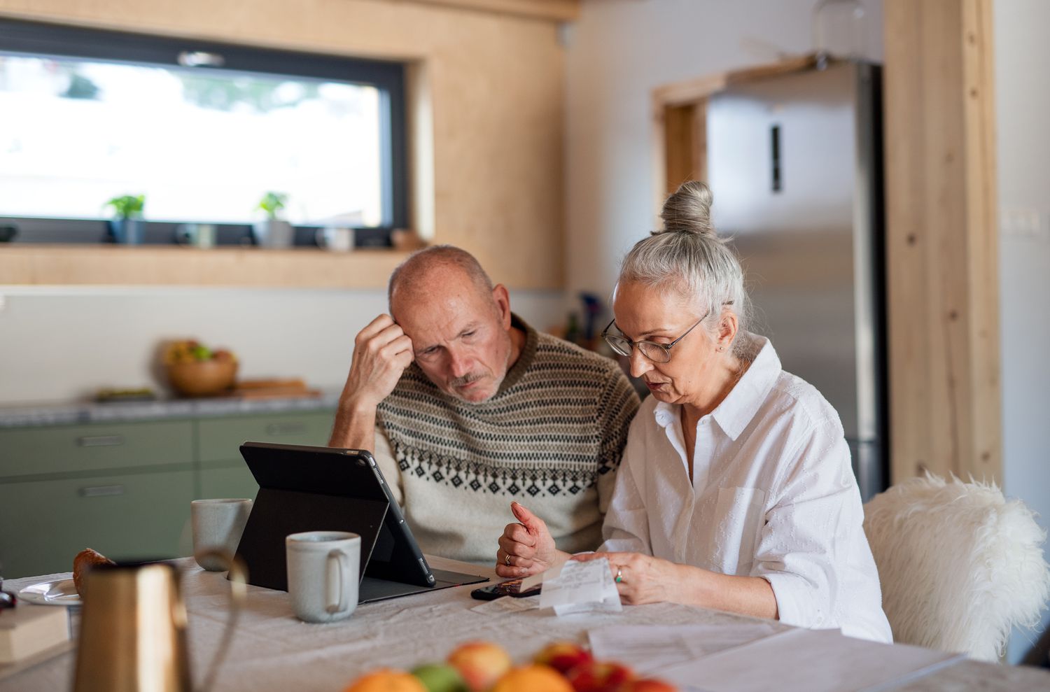 An older couple sits at the kitchen table with croissant and cups of tea, and together they look at a digital tablet screen and paper receipts, perhaps evaluating their budget.