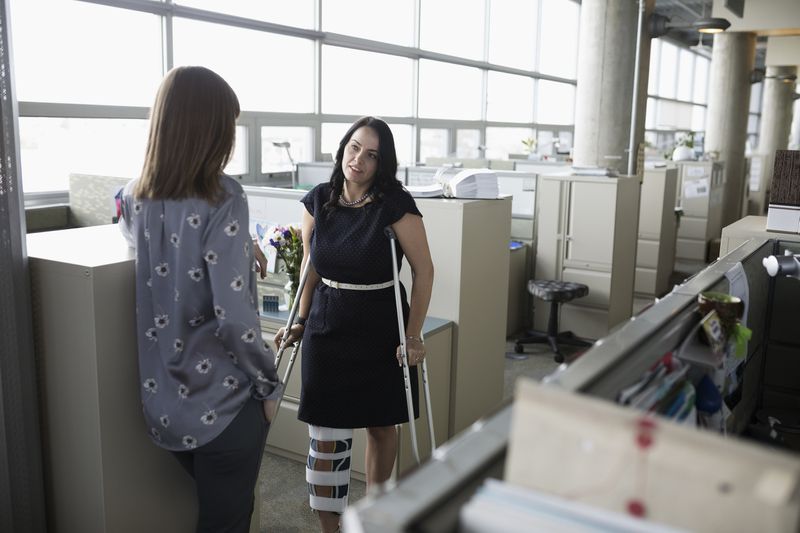 Businesswoman in leg brace on crutches talking to colleague in office