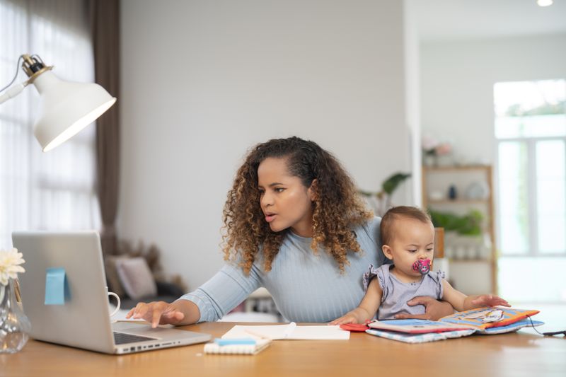 Young mom sitting at her kitchen table, looking at a laptop while holding a toddler on her lap.