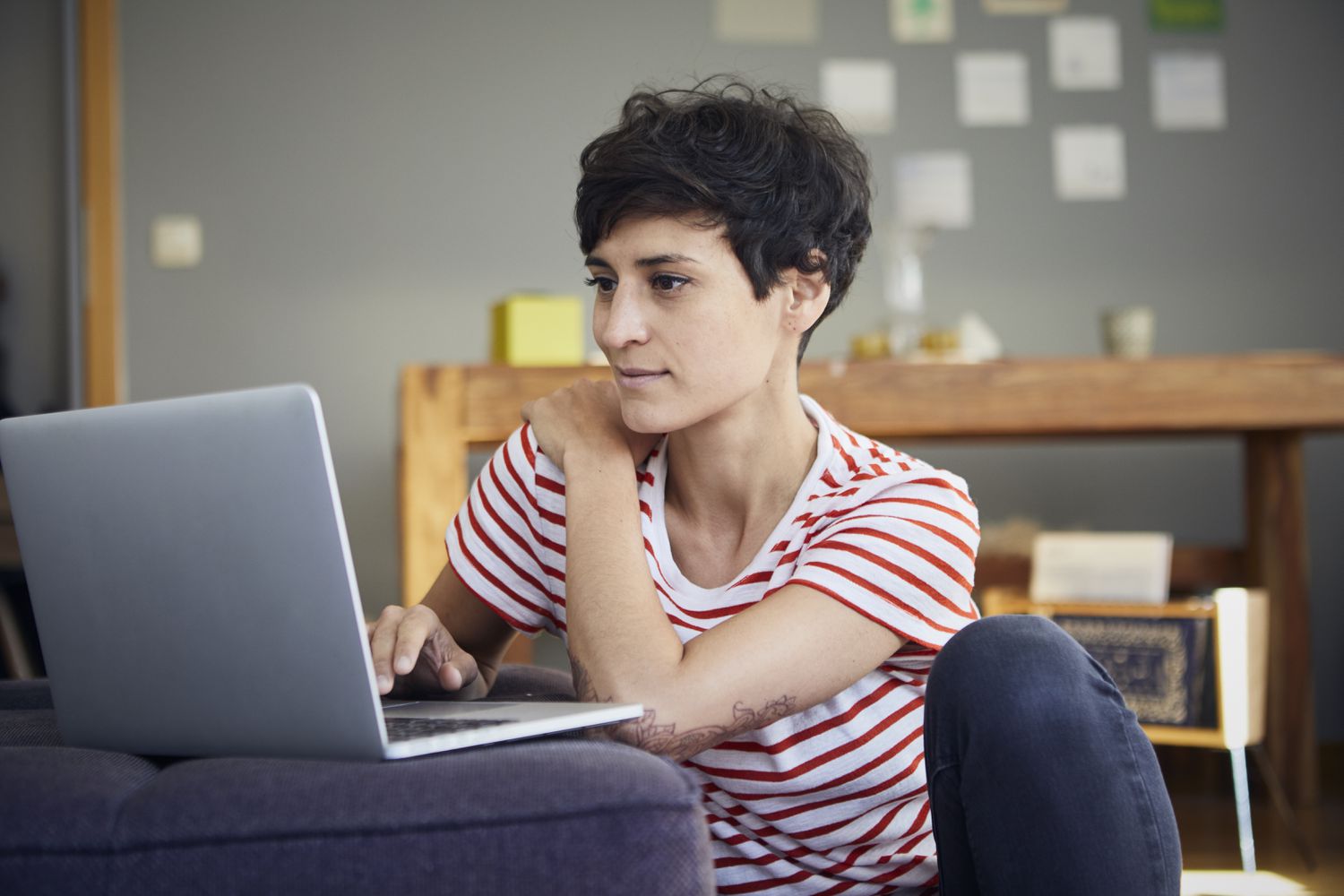 A woman in a red and white striped t-shirt sits on the floor against a couch with a laptop