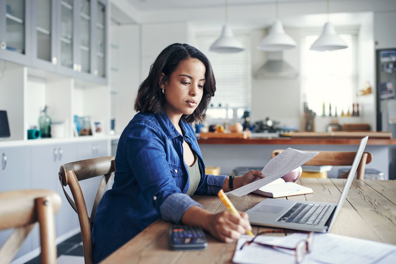 A woman reviews paperwork and uses a laptop in her kitchen