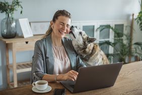Woman working on her laptop, interrupted by her dog who licks her face.