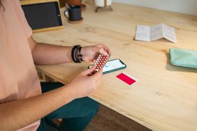 Woman holds birth control pills near her cell phone while resting her hands on a desk.