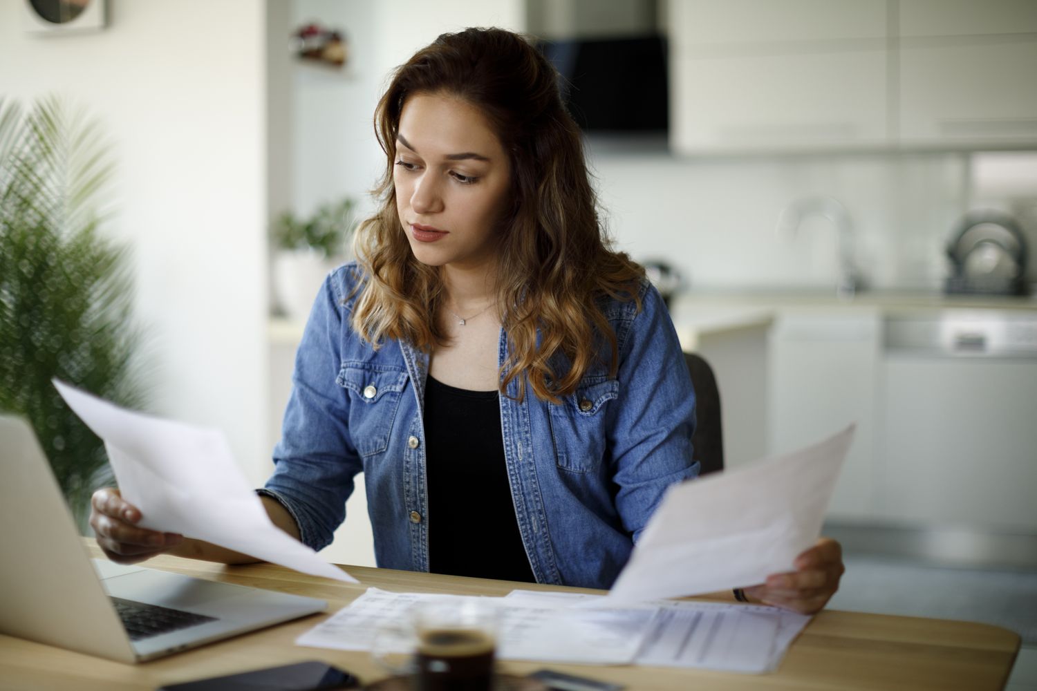 A woman sits at a table looking at documents in front of a laptop