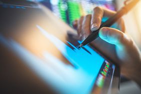 a hand holding a stylus used on a tablet with a chart with stock tickers in the background