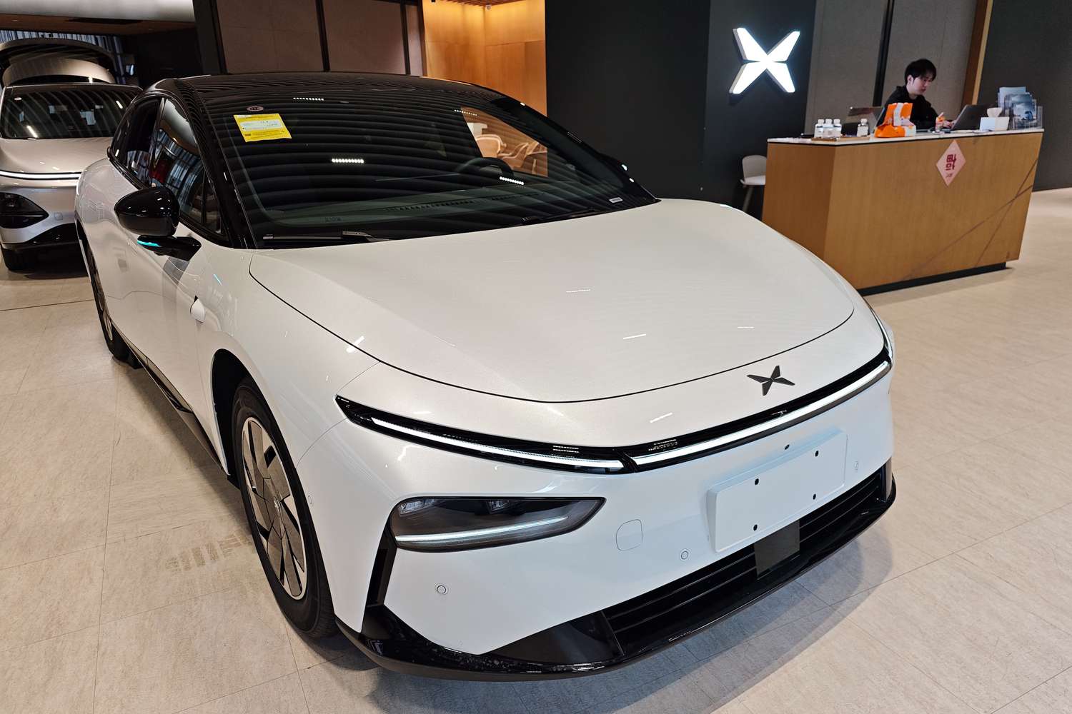 A white XPeng G6 car in a showroom, with a person sitting behind a desk to the right of the car