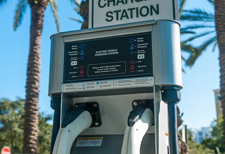 Electric vehicle charging station sign on top of a public charging station with palm trees in background.