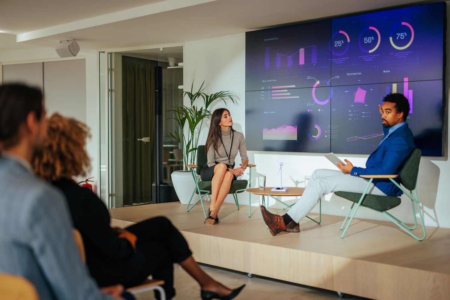 Two company representives sit on stage before a large display of company data to hold a press conference about the company's activities.