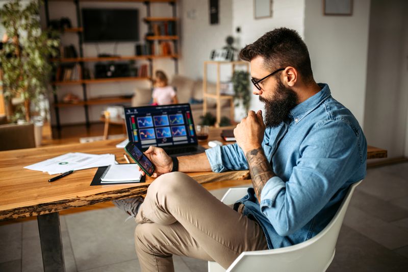 Mid-adult man working at home using laptop, while his daughter doing her homework in background in living room.