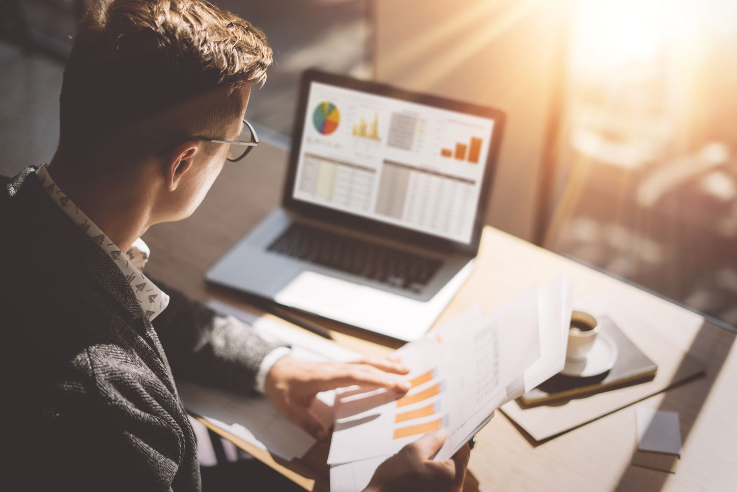 Young market analyst looking at laptop and papers with financial information and an espresso on his desk
