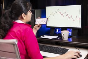 Young woman looking at charts on a monitor and a cell phone.