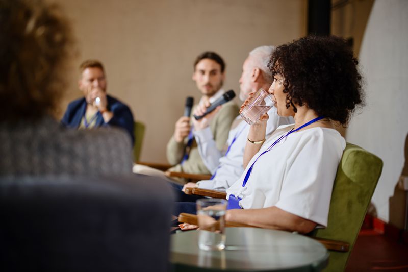 Panelists sitting, woman is drinking and two are holding microphones.