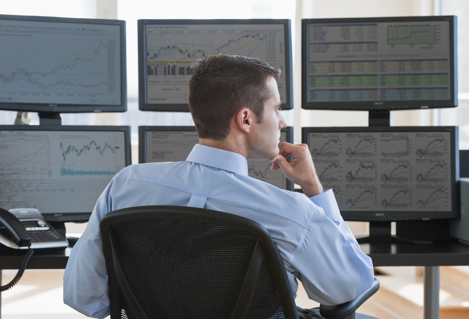 A broker-dealer sits at their desk and reviews financial information displayed on several computer monitors in front of them. 