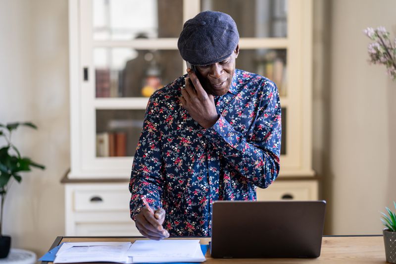 Man standing at a desk talking on a mobile phone with an open. laptop in front of him