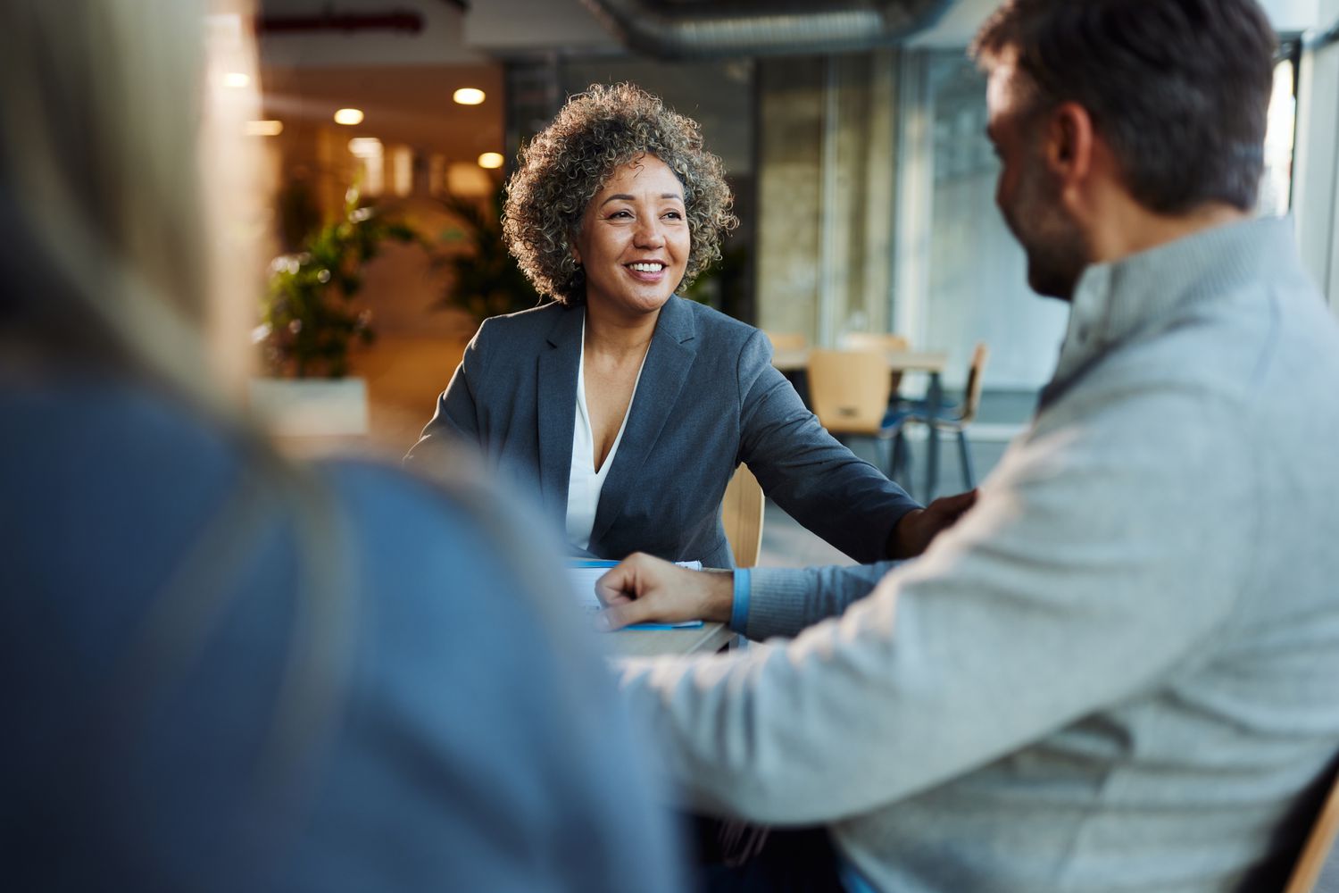 A woman wearing a suit sits a table and talks to two other people, who have their backs to the camera.