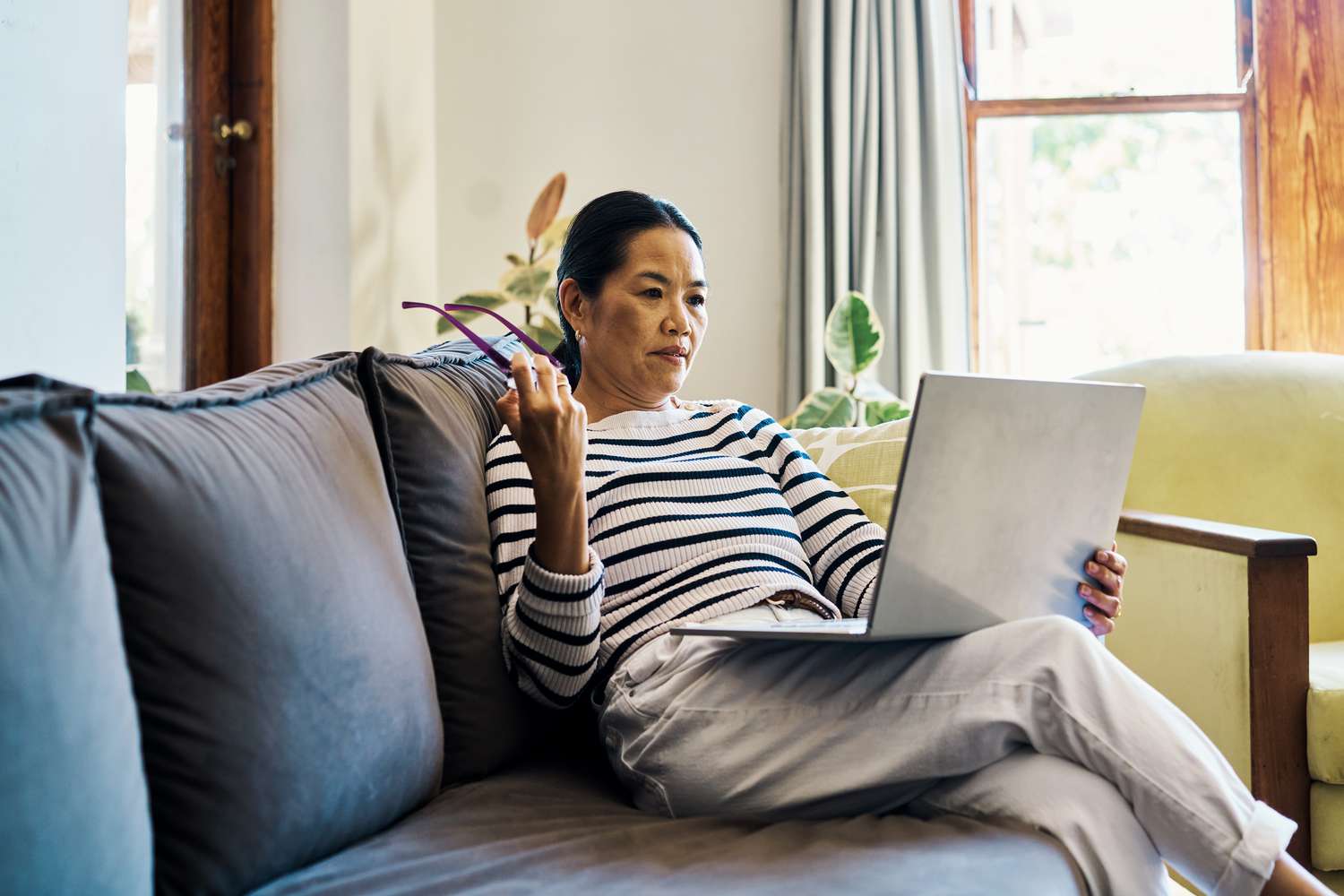 A woman sits on a couch with her laptop on her lap holding glasses in her right hand