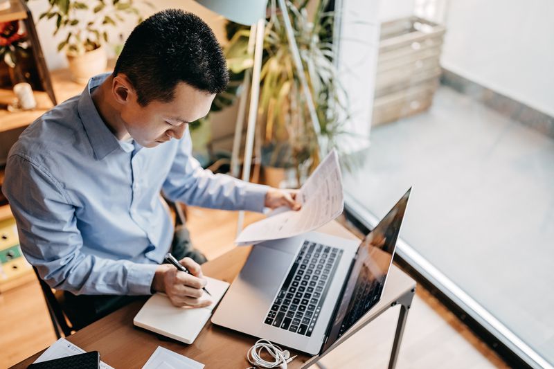 A young man calculates his home expenses, sitting at table with a laptop, a pen, and papers in hand
