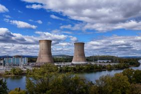 A pair of nuclear reactors are seen in above a river and trees, with cloudy blue sky in the background.