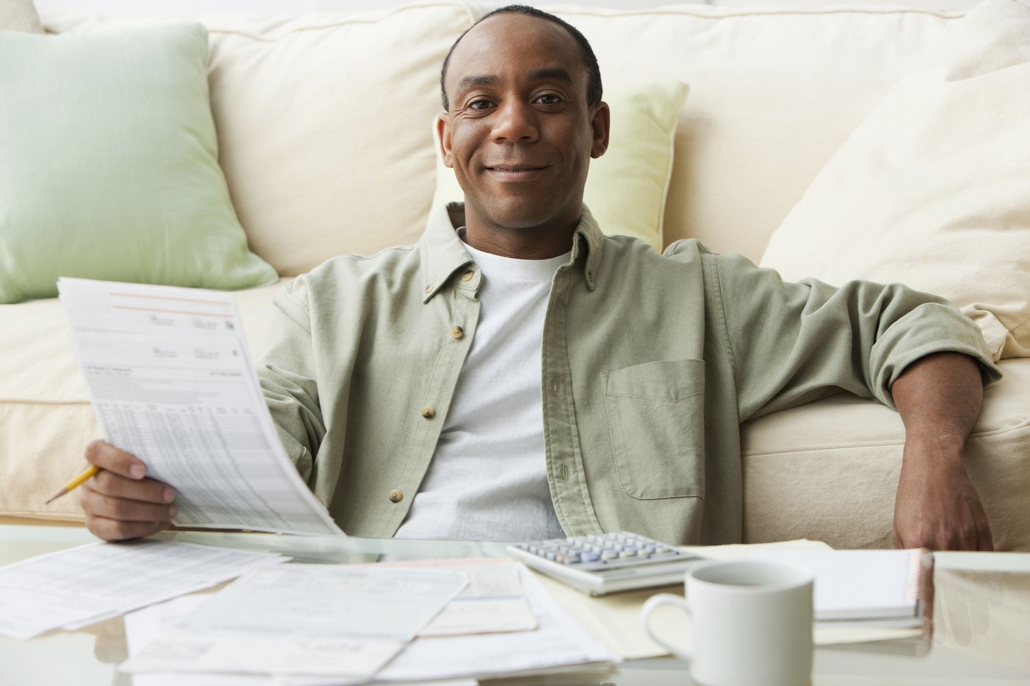 A man paying bills at his coffee table with papers, a calculator, and a coffee mug on the table