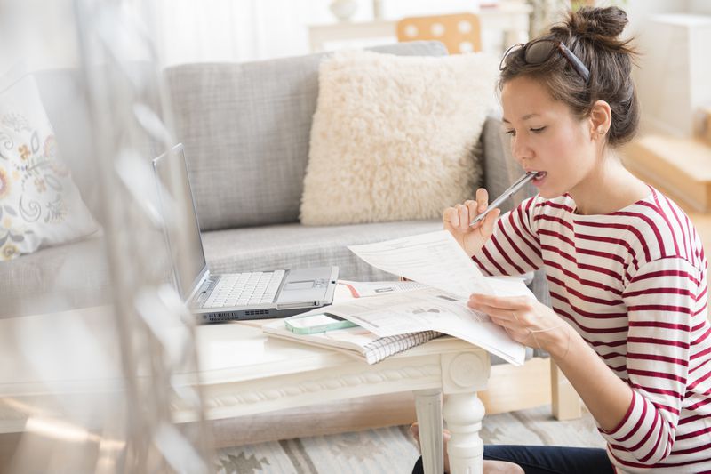 A young woman with her hair tied up and sunglasses perched on her head sits on the floor near a low table in a living room. She is wearing a red and white striped shirt and holding a pen to her mouth while concentrating on papers in her hands. A laptop, notebook, and other documents are spread out on the table in front of her, suggesting she is working or studying. The cozy room features a gray couch with decorative pillows and a fluffy beige cushion in the background.