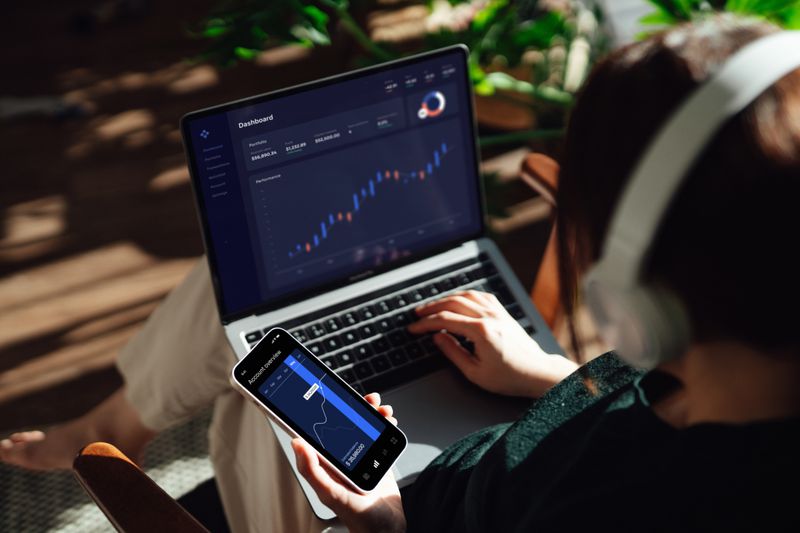 Person with headphones on laptop and phone tracking investments and trading stocks.