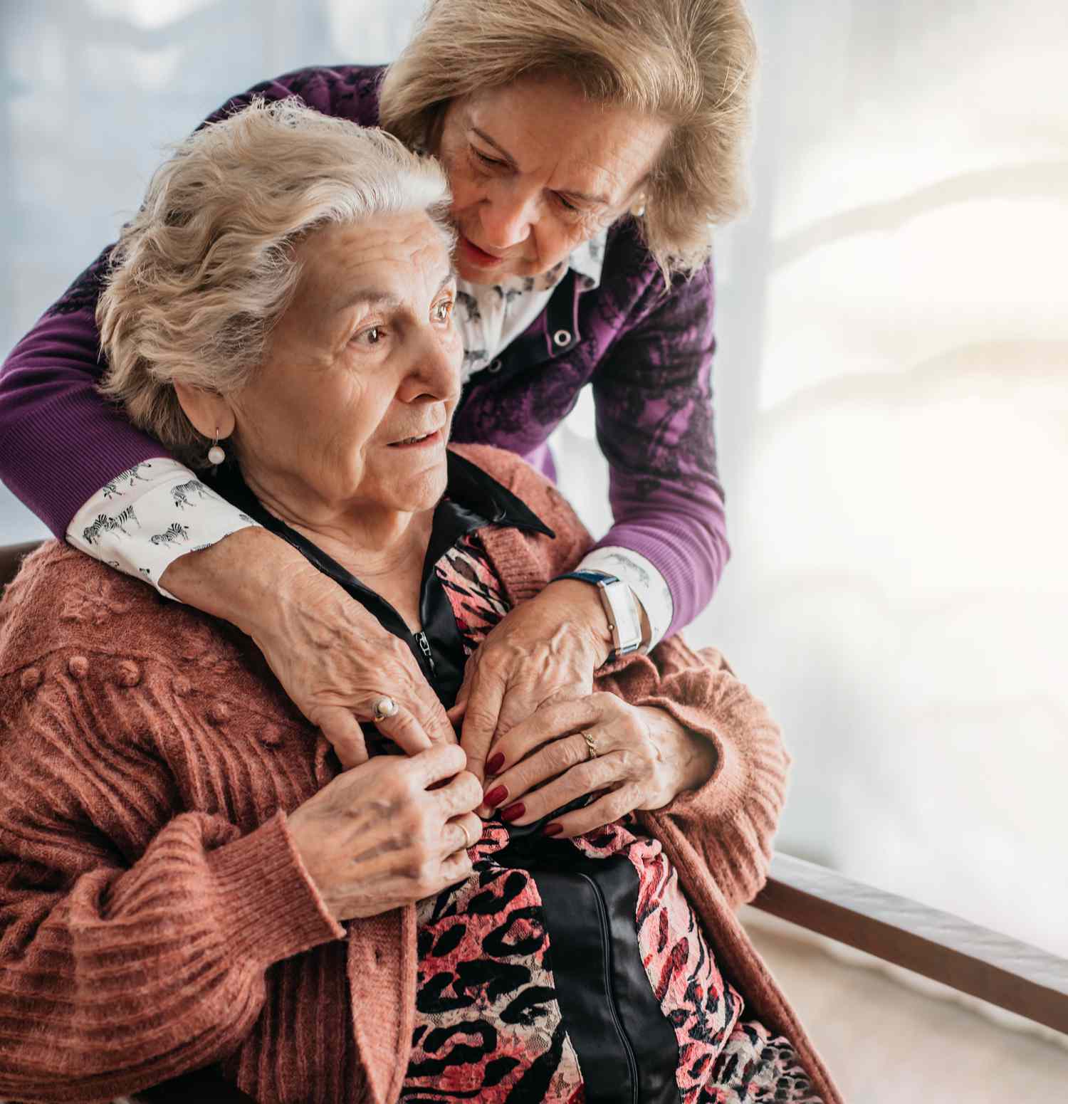 An adult daughter helps their mother adjust a sweater as they assist nursing home staff administering care.