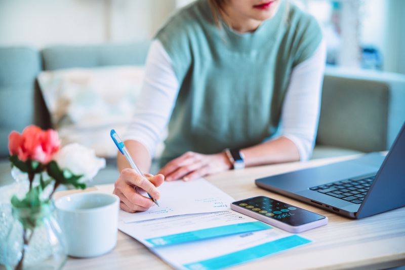 A person looking at a computer, pen in hand writing notes a sheet of paper.