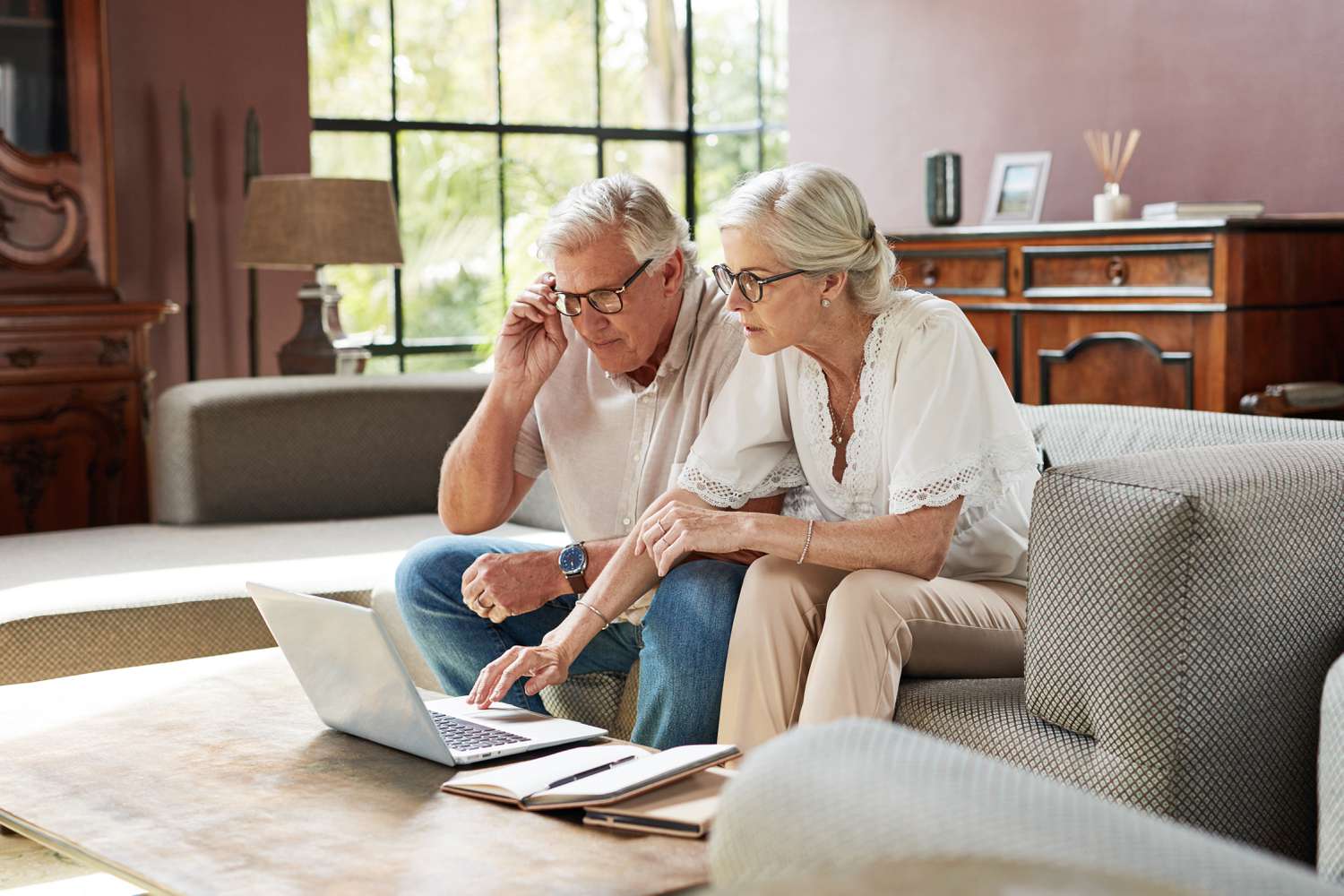 An older couple sits next to each other on a couch, looking at a laptop computer.