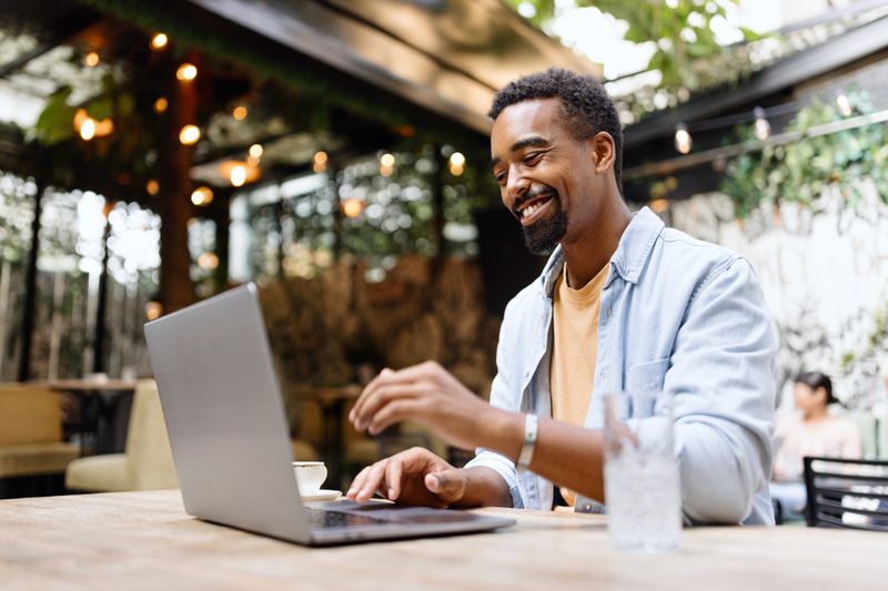 A happy man sits in a cafe using a laptop with a big smile on his face.