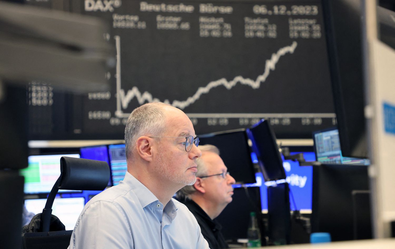 Two analysts look at computer screens displaying the closing price of stocks in a brokers office surrounded by screens showing other stock and index data.