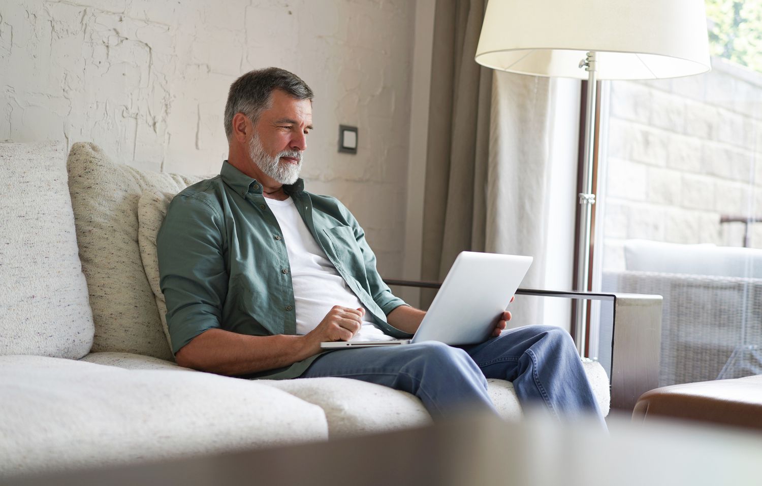 Man in his 50s or 60s sitting on his living room couch and studying something on his laptop