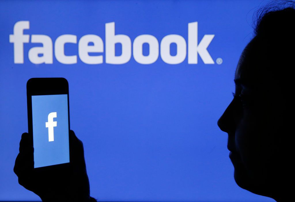 The Facebook logo is displayed on the screen of an iPhone in front of a TV screen displaying the Facebook logo.
