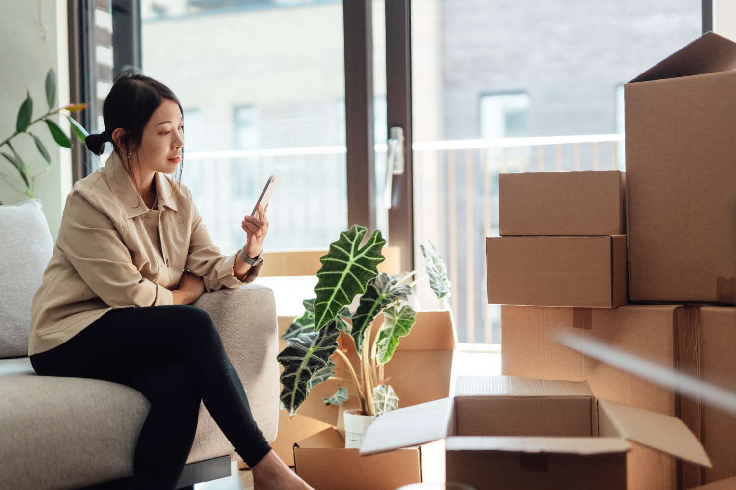 A woman sits on a couch in a living room with packing boxes looking at her smartphone