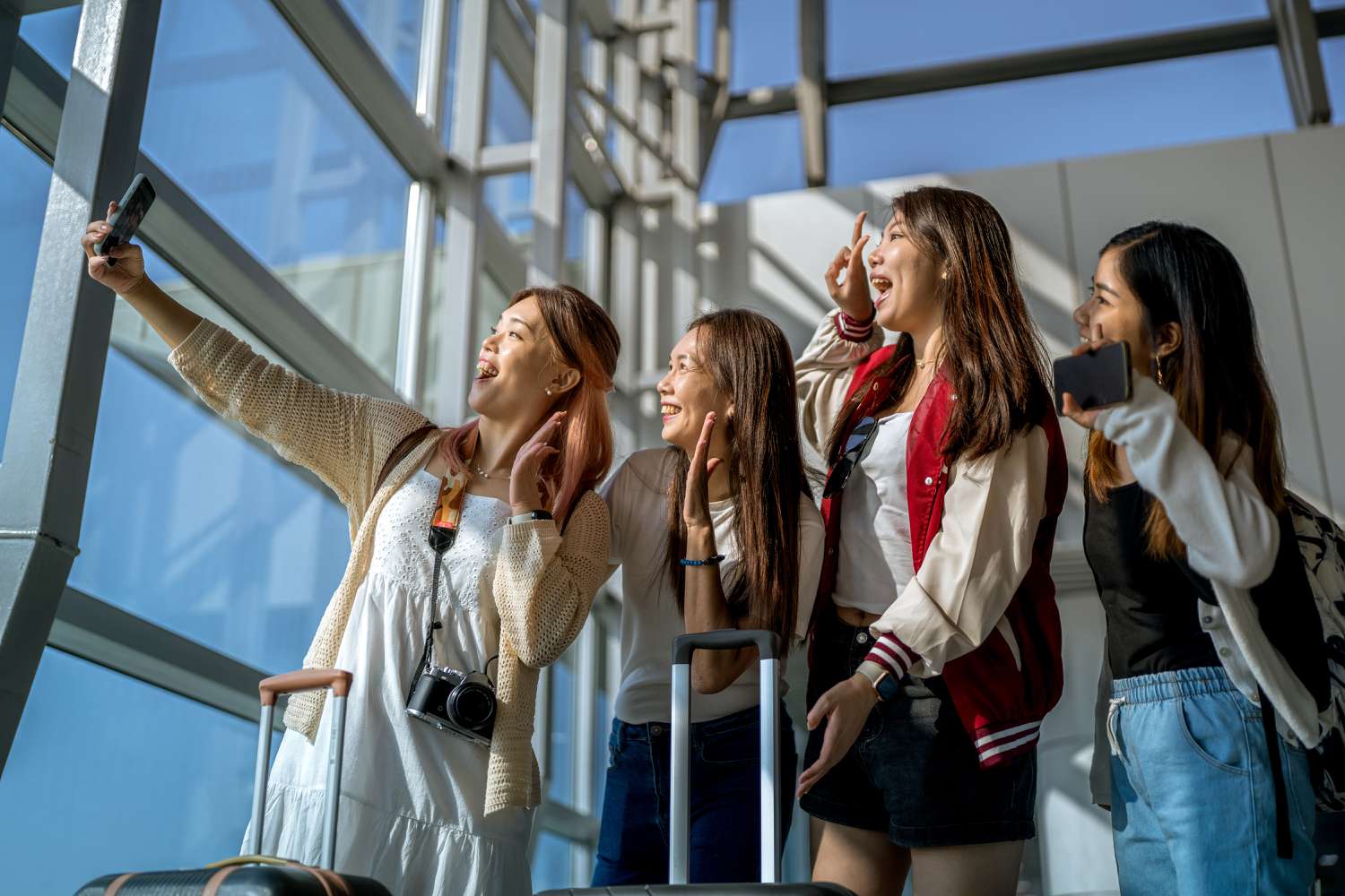 A group of women in an airport with luggage, taking a selfie in front of a group of windows. 