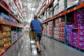 A man with his back to the camera pushes a shopping cart down an aisle at Costco.