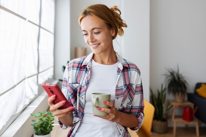 Woman in her 20s or 30s at home holding a coffee cup and looking happily at something on her smartphone
