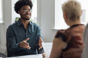 Over the shoulder view of a young male employee engaging in a focused discussion with a mature female manager at the managers desk in an office.