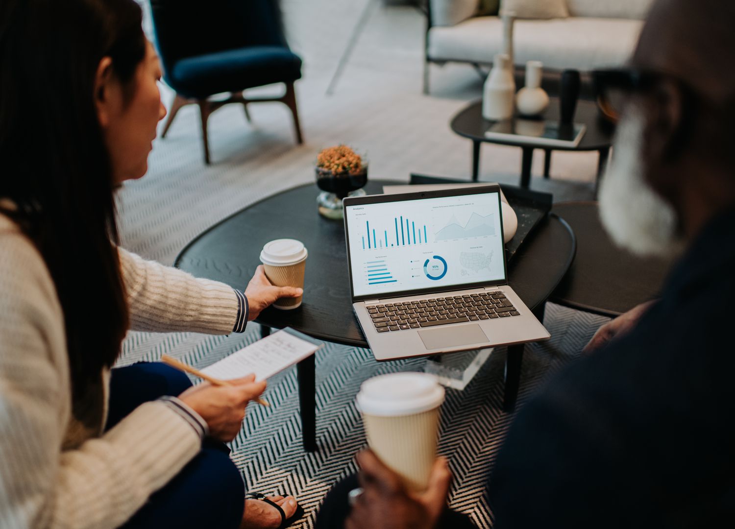 Two people sit on a sofa. They share a laptop, both looking at the statistics displayed, while they drink coffee.