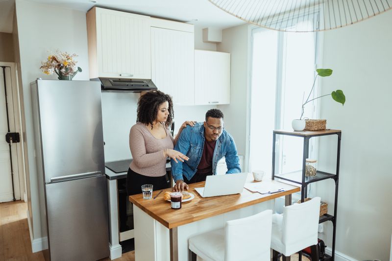 A couple standing together in a well-lit kitchen discuss something on a laptop screen.