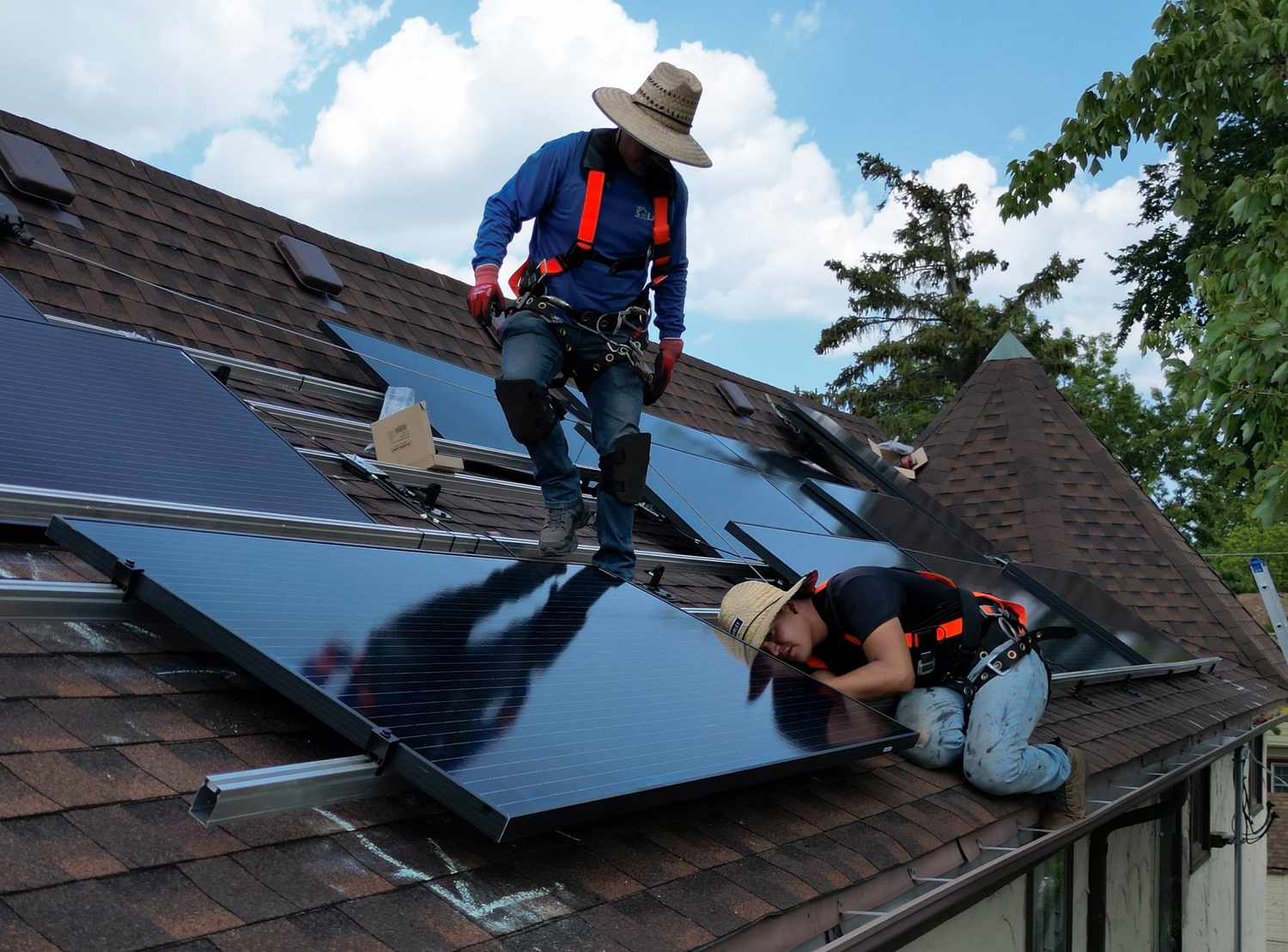 Two workers install solar panels on the roof of a home next to trees and a blue sky.