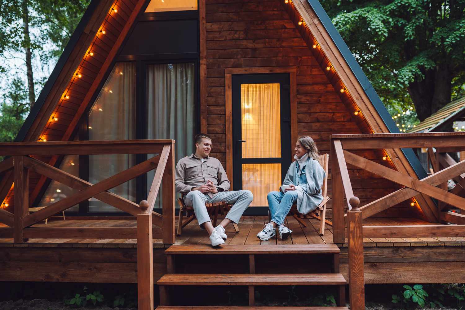 A man and a woman sit on a deck of a triangular cabin in the woods