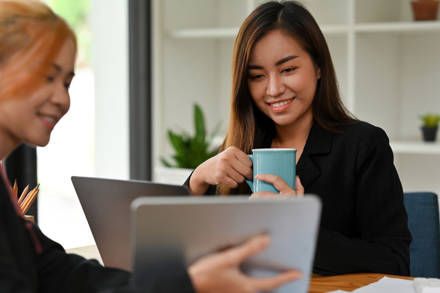 A woman sits at a table, looking at a tablet computer held by someone else and holding a coffee mug.