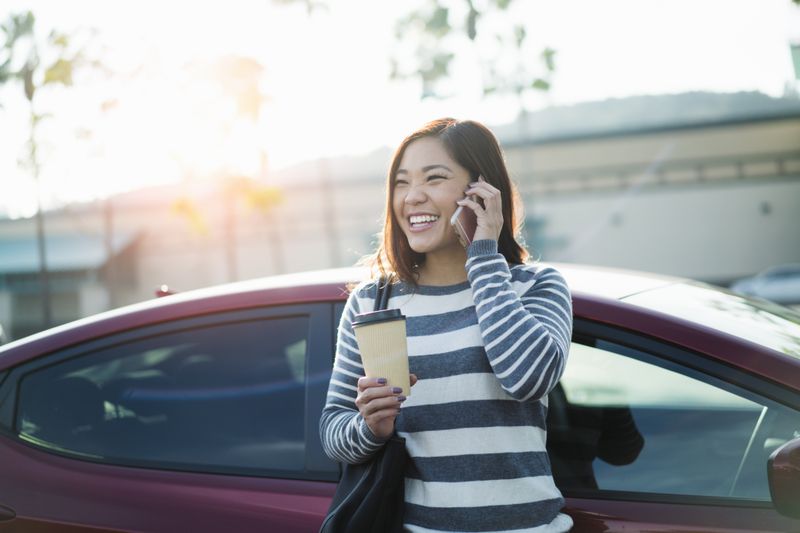 Woman on phone with coffee in front of car.