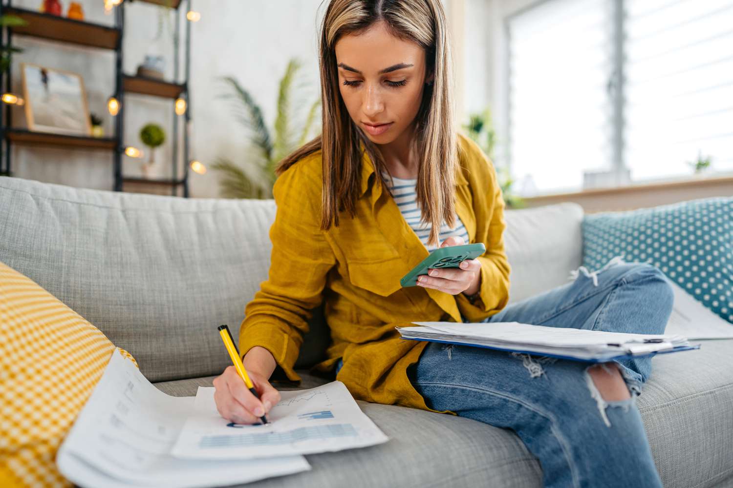 Young person with long hair sits on a gray couch reviewing their finances on their smartphone. 