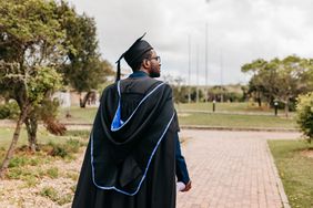 Young male graduate with beard wearing graduation gown and mortarboard walking and looking sideways into distance on campus grounds