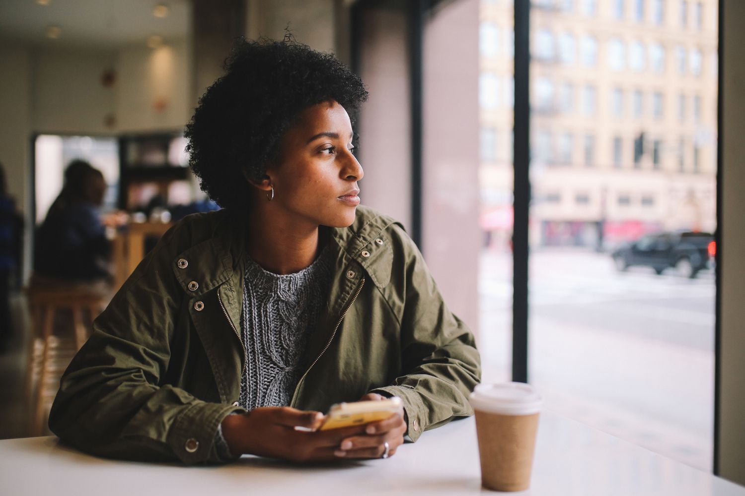 A woman sits at a cafe with a smartphone in hand, looking out the window