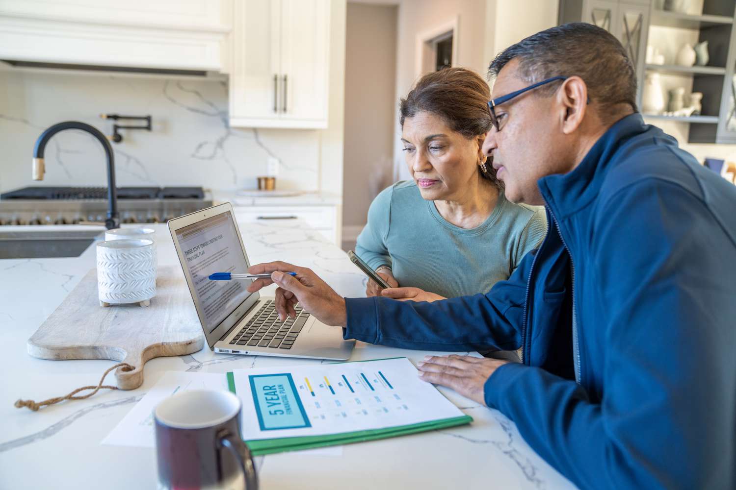 A couple sit at their kitchen island with a laptop open and their bills spread out in front of them as they review their finances.